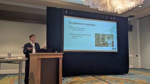 Photo of Speaker at podium next to large screen showing presentation slide.