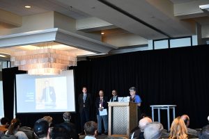 A person is at a podium with three other people on stage next to the podium.