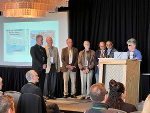 A person at a podium with 6 other people on stage next to the podium.