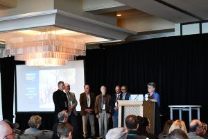 A person at a podium with 6 other people on stage next to the podium.