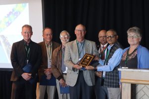 A person receiving an award plaque, and surrounded by colleagues.