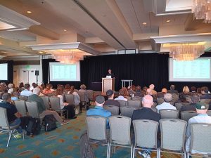 Conference attendees seated in theater style rows with speaker at a podium