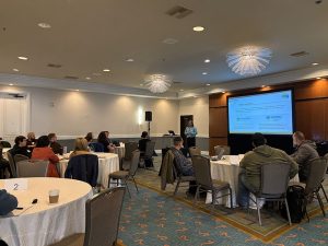 Speaker next to screen with presentation to group of attendees seated at tables.