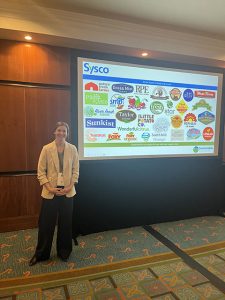 Photo of a young lady by a screen with projected logos exhibiting brands owned by Sysco.